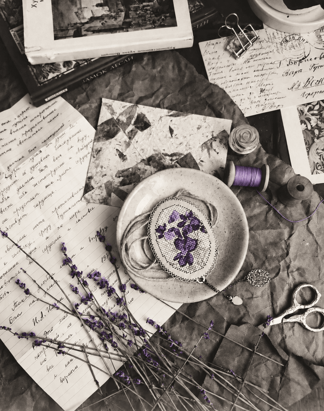Sewing materials and handwritten papers in a softly aged photograph, with violet cross-stitch, thread and lavender petals